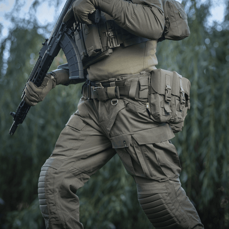Henkilö pukeutuneena M-Tac War Belt ARMORiin ja taktiikka-varusteisiin luonnossa, valmis taistelutehtäviin.