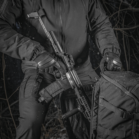 M-Tac Rygsæk Gen.II Elite Large, henkilö pitää asetta repun vieressä metsässä; repun kestävä muotoilu näkyy.