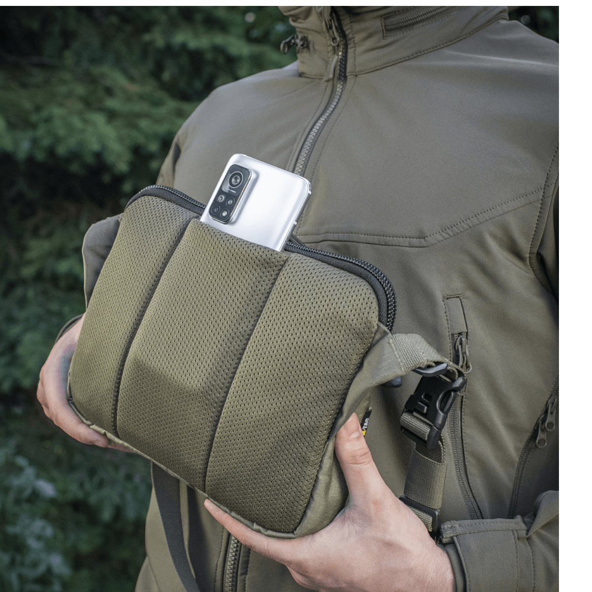 M-Tac Admin Taske Elite Cordura-kankaasta, älypuhelin taskussa, säädettävä olkahihna ja tukeva rakenne, ulkoilmataustalla.