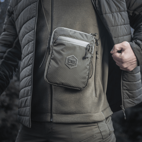 M‑Tac Pocket Bag Elite HEX kannetaan miehen edessä ulkovaatteissa, näyttää laukun kompaktin ja vankan muotoilun YKK-vetoketjuilla.