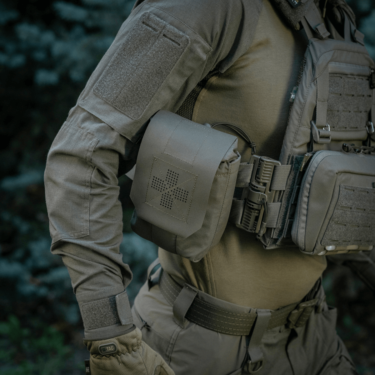 M-Tac Medic Pouch Vertical Large Elite kiinnitettynä taktiseen liiviin metsässä, tarjoaa kestävän säilytyksen ensiapuvälineille.