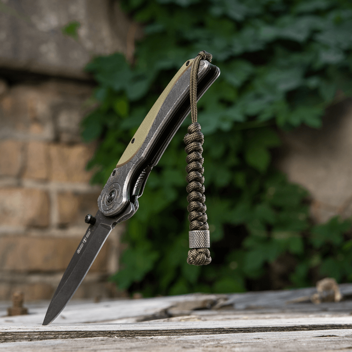 M-Tac Viper Lanyard Stainless Steel ruostumattomalla helmellä ja paracordilla veitsen vieressä ulkoilmataustalla.