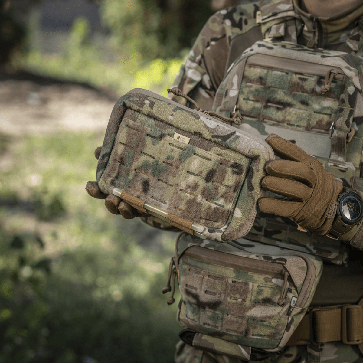 M-Tac Admin Pouch Large Elite kamuflaasiasuiseen henkilöön tarttuneena, näyttää kestävän ja toiminnallisen muotoilun taktiseen käyttöön velcro-paneeleilla.