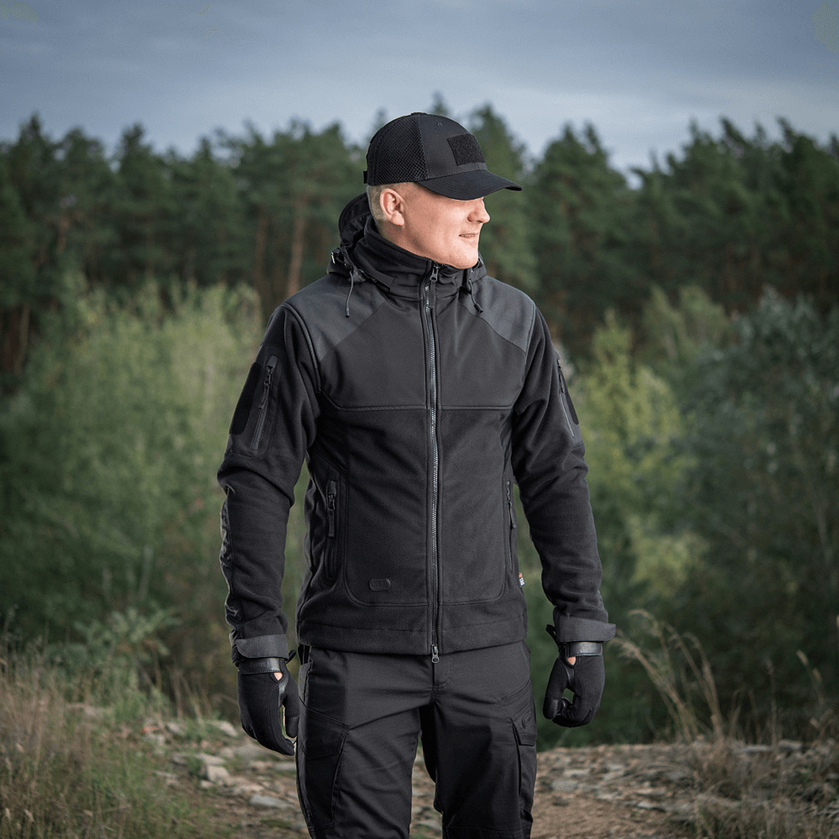 Mies M-Tac Norman Windblock -fleecetakissa metsäalueella, sopii kylmään ja tuuliseen säähän, näyttää takin toiminnallisuuden ja muotoilun.