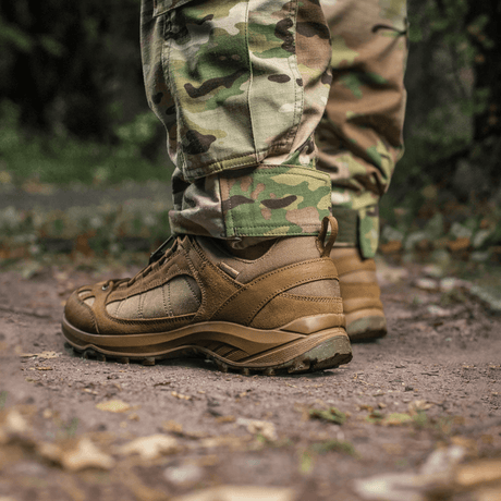 Henkilö pukeutuneena M-Tac-housuihin STURM NyCo Extreme Gen.II ja taktisiin saappaisiin metsäpolulla, sivulta nähtynä.