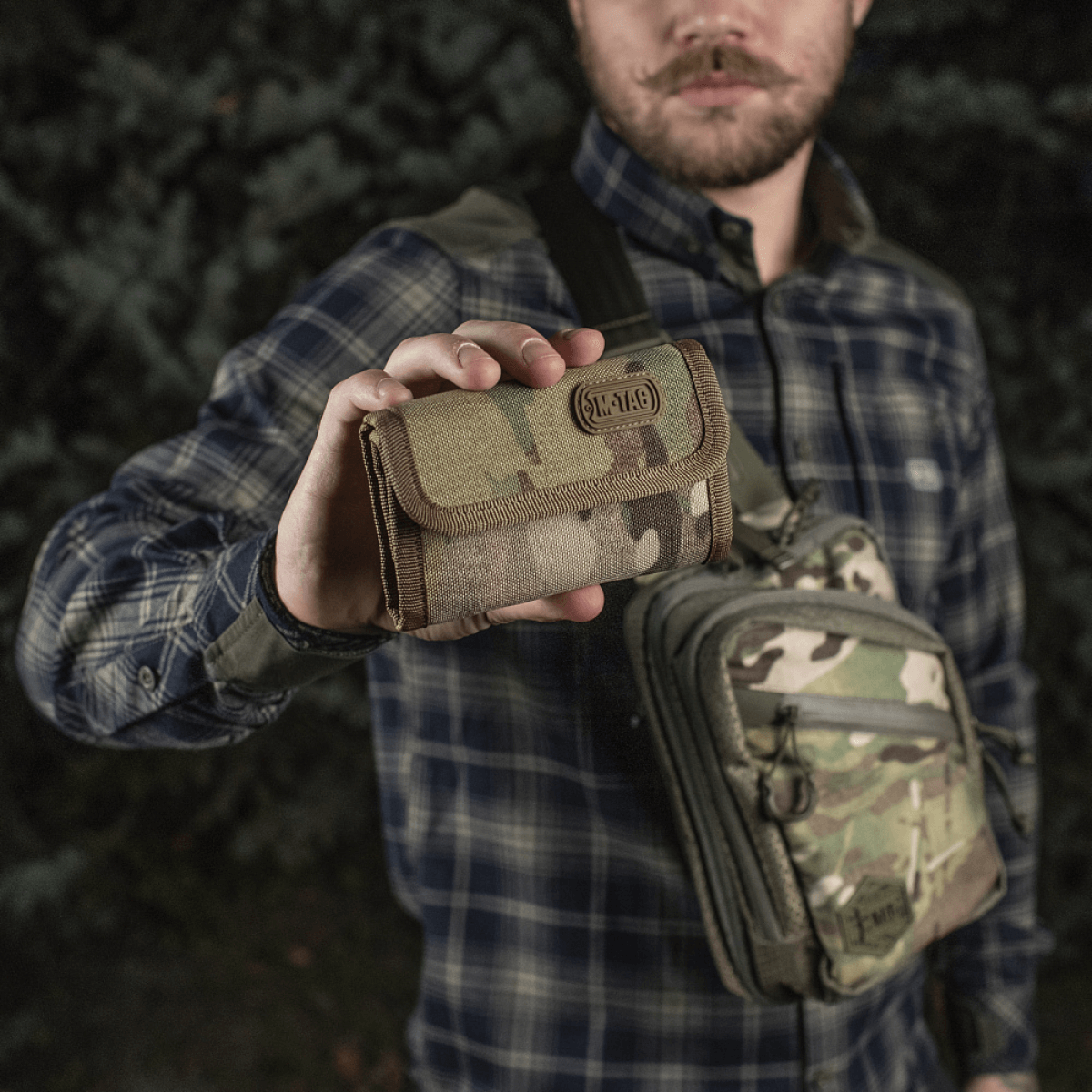 Mies pitelee M-Tac Pung Elite Gen.II:tä maastokuviossa, valmistettu kestävästä Cordura 500D:stä, käytännölliset lokeroita ja YKK-vetoketjut.