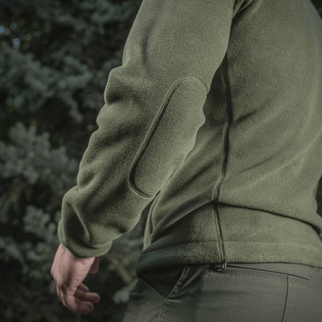 Mies pukeutuneena M-Tac Nord Polartec -fleecetakkiin vihreänä, suunniteltu tyyliin ja toimivuuteen, nähtynä takaa ulkoilmassa puiden ympäröimänä.