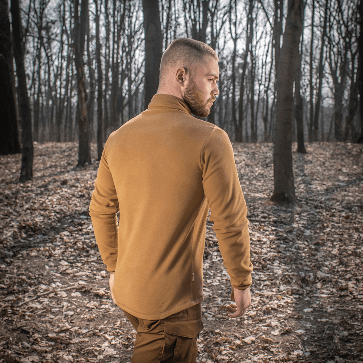 Mies pukeutuneena M-Tac Delta Polartec fleece-takkiin metsässä nähtynä takaa, näyttää tyylin ja toiminnallisuuden luonnoympäristössä.
