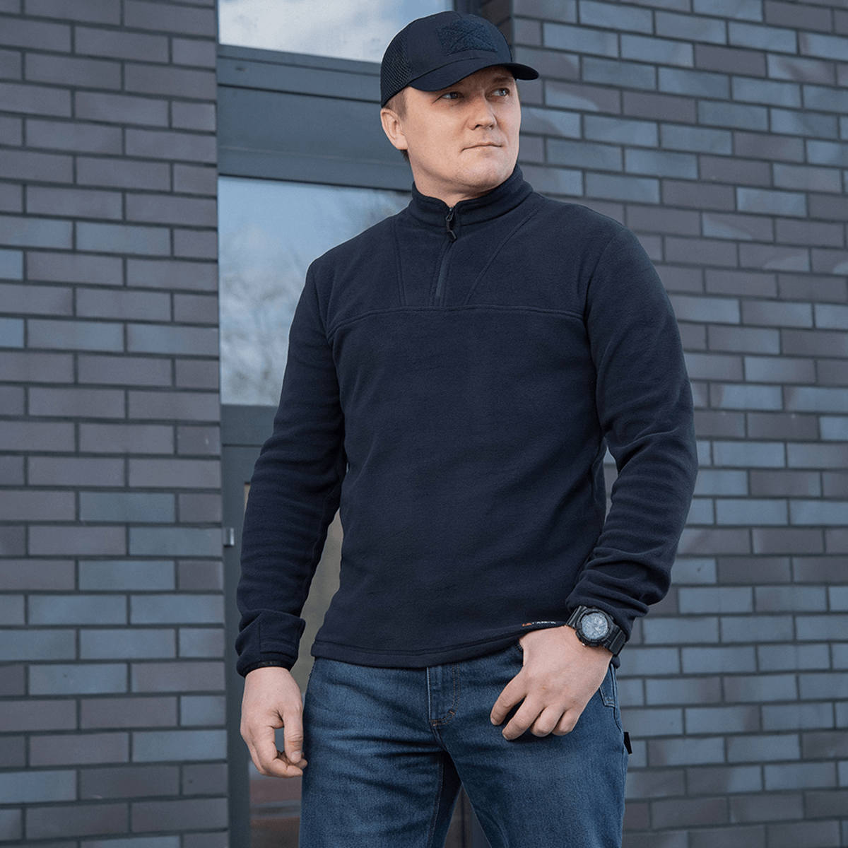 Mies pukeutuneena M-Tac Delta Polartec -fleece-takkiin muuriseinän edessä.