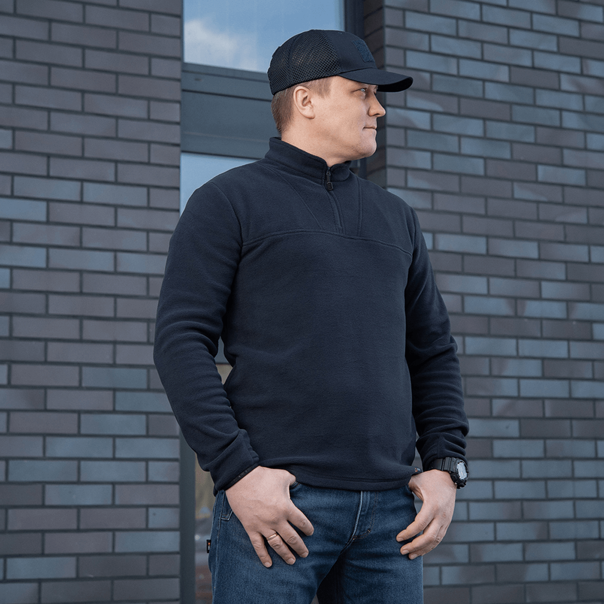 Mies käyttää mustaa M-Tac Delta Polartec -fleece-takkia Polartec®-fleecestä muuriseinän edessä.