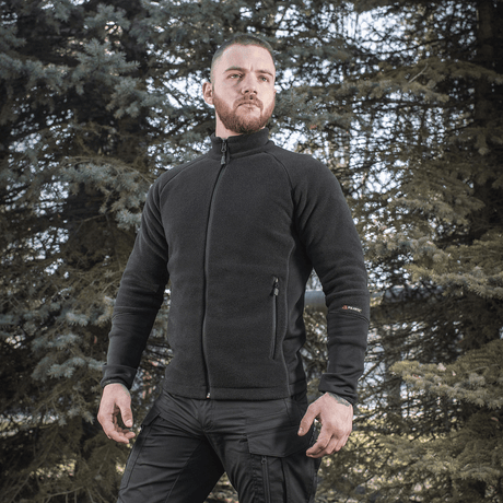Mies pukeutuneena M-Tac Polartec Sport -fleecatakkiin mustana, sopii vaihtokauteen ja viileään säähän, ympäröity puita.