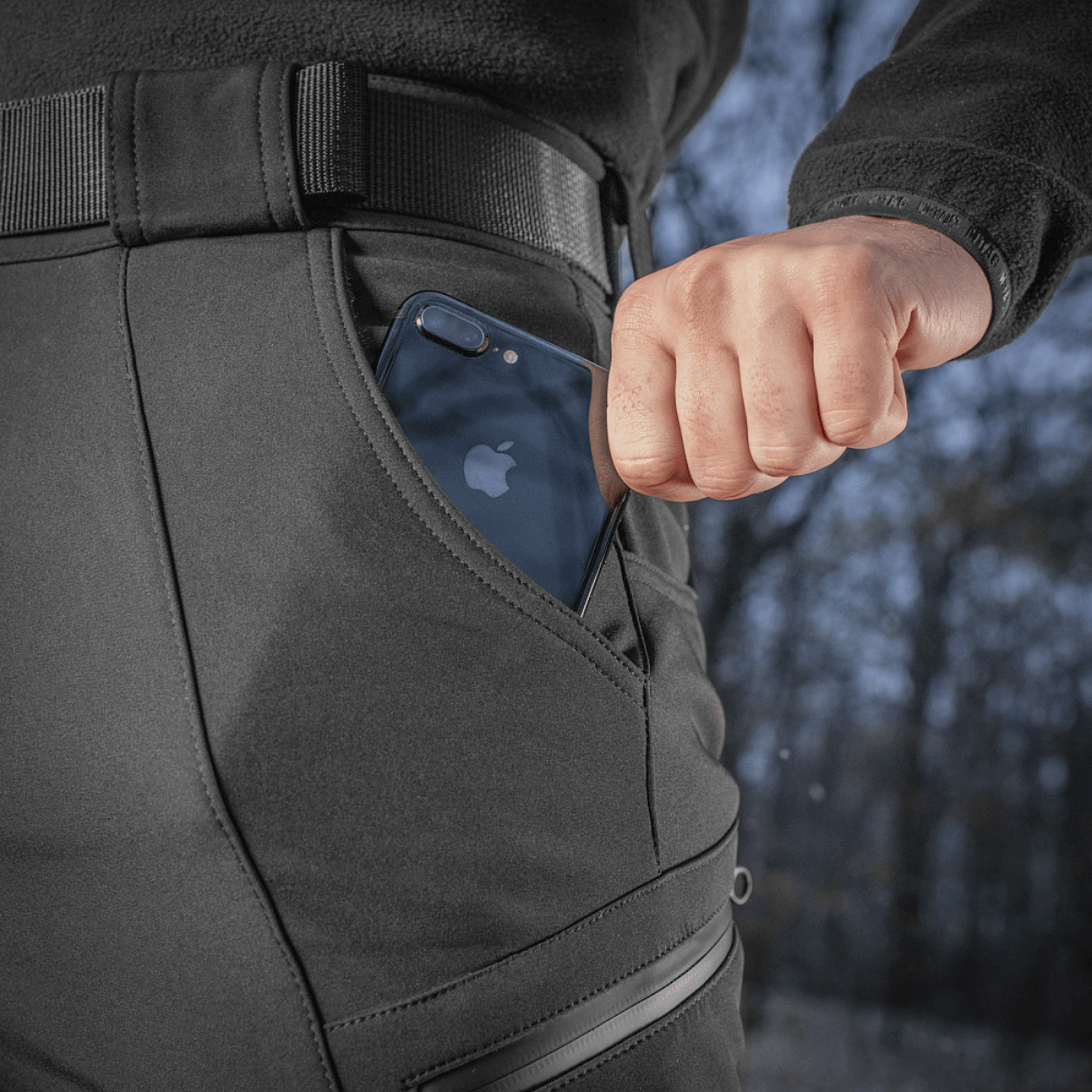 Henkilö pukeutuneena M-Tac Soft Shell Vinter -housuihin, puhelin taskussa, korostaa käytännöllistä ja toiminnallista muotoilua kylmiin oloihin.