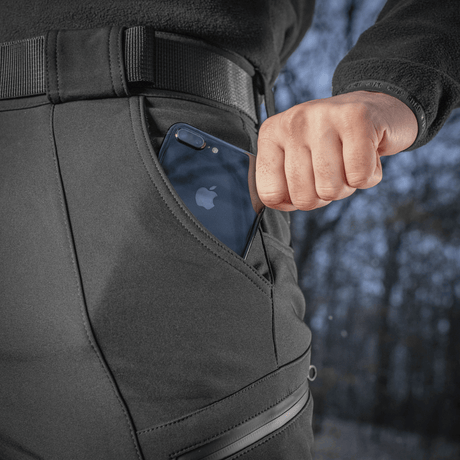 Henkilö pukeutuneena M-Tac Soft Shell Vinter -housuihin, puhelin taskussa, korostaa käytännöllistä ja toiminnallista muotoilua kylmiin oloihin.