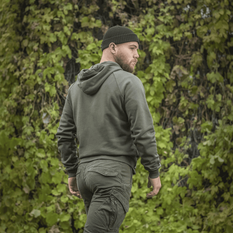 Mies M-Tac Cotton Raglan -hupparissa vihreiden lehtien edessä, näyttää raglanhihat ja hupun, mukava vaatetus.