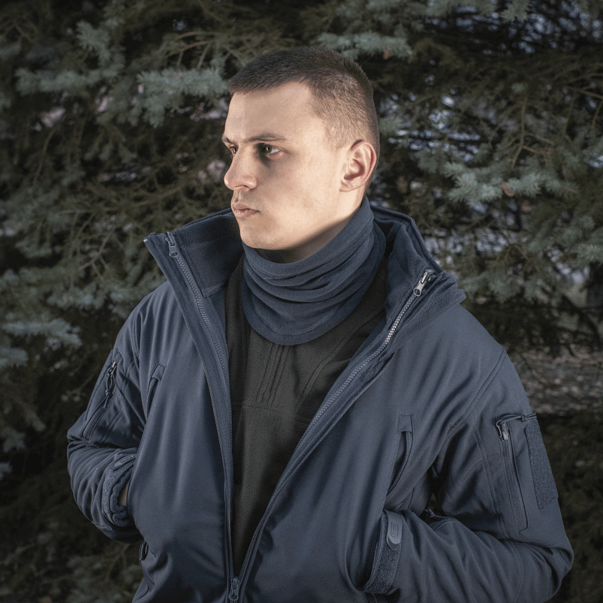 Mies sinisessä takissa ja M-Tac Anatominen Tube Scarf Polartec-nyörillä seisoo puun edessä, suosittu talvikäyttöön.