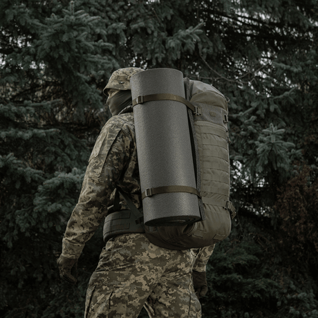 Hahmo camo-vaatteissa kantaa reppua, jossa M-Tac Liggeunderlag 20 mm (60x180 cm), metsäympäristössä.