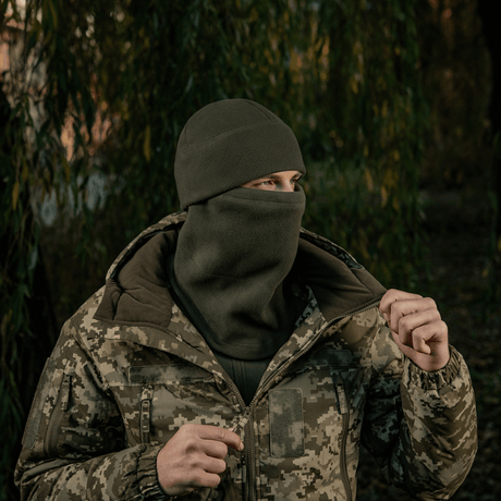 M-Tac Anatomisk Snood Justerbar Elite Fleece (320 g/m²) pidetään balaclavana kylmässä säässä, taustalla metsä ja maastokuvioinen takki.