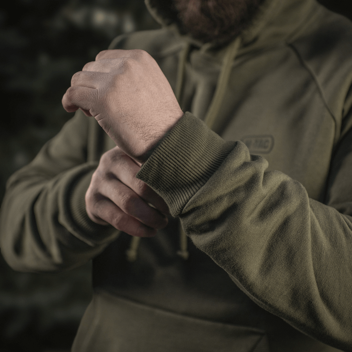 Henkilö pukeutuneena M-Tac Cotton Raglan Hard Hoodieen 100% puuvillaa fleece-vuorella vihreänä, näyttää joustavan hihansuun, sopii viileämpiin vuodenaikoihin.