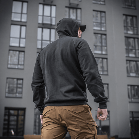 Mies M-Tac Cotton Raglan Hard Hoodiessa, 100% puuvillaa fleece-vuorella, sopii syksyyn ja talveen, nähty takaa rakennuksen edessä.