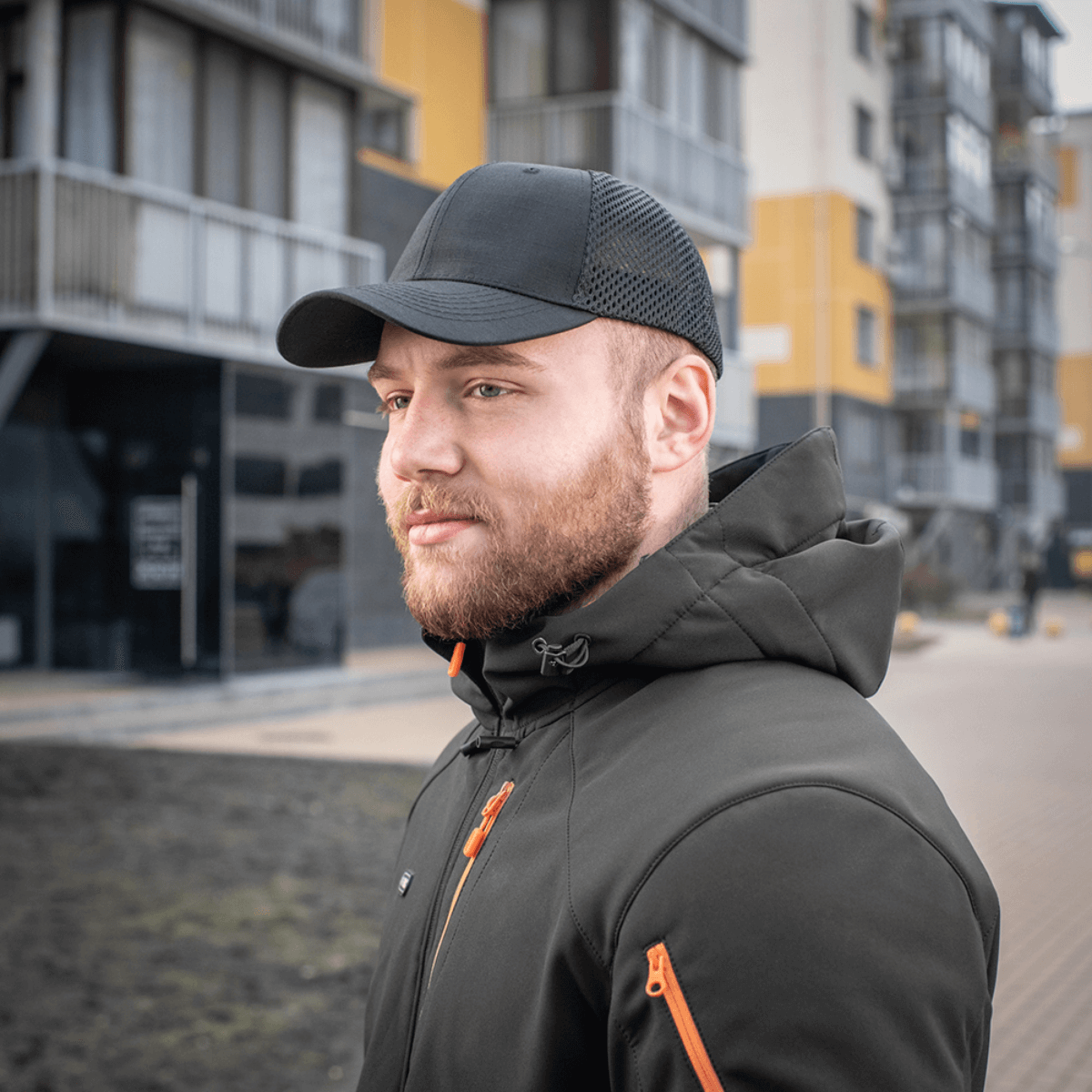 Miehellä on M-Tac Mesh Flex Ripstop Baseball Cap modernien rakennusten edessä, täydellinen ulkoiluun, musta lippis mesh-paneeleilla.