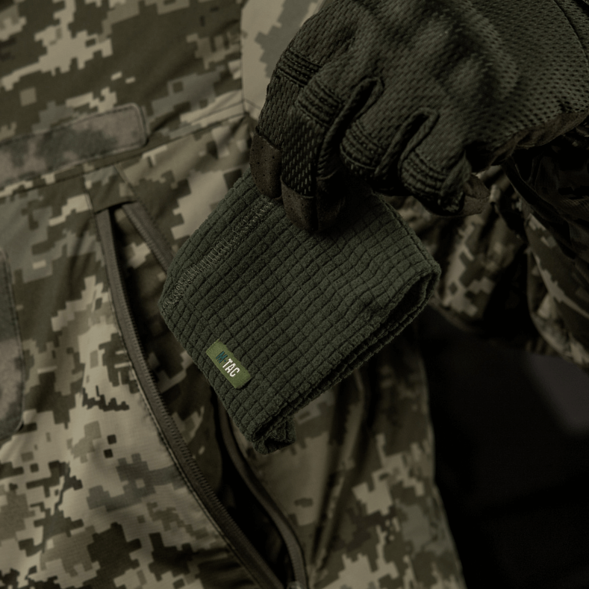M-Tac Rip-Stop Fleece Hjelmliner – lämmin ja kestävä fleece-kypäränvuori, pidetään kädessä, täydellinen viileään säähän, maastokuvioista taustaa vasten.