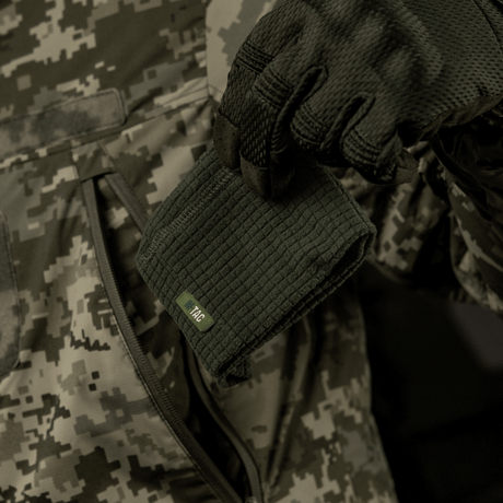 M-Tac Rip-Stop Fleece Hjelmliner – lämmin ja kestävä fleece-kypäränvuori, pidetään kädessä, täydellinen viileään säähän, maastokuvioista taustaa vasten.