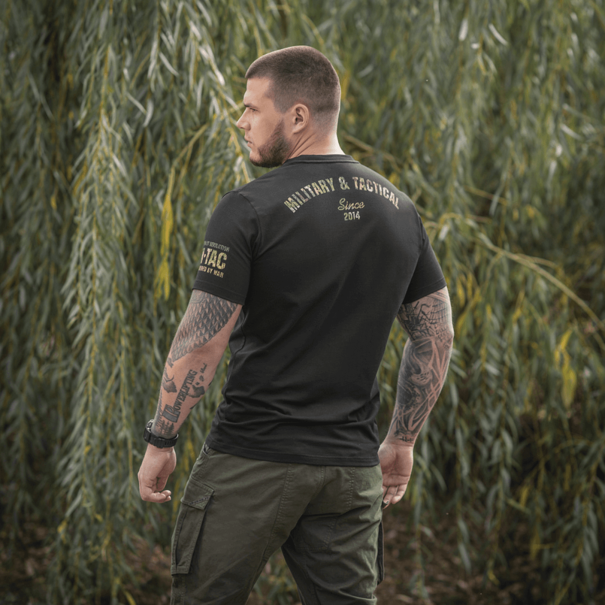 M-Tac T-Shirt Logo sotilaallisella camokuvioinnilla ja sloganilla "Born by Revolution – Hardened by War" luonnon taustalla.