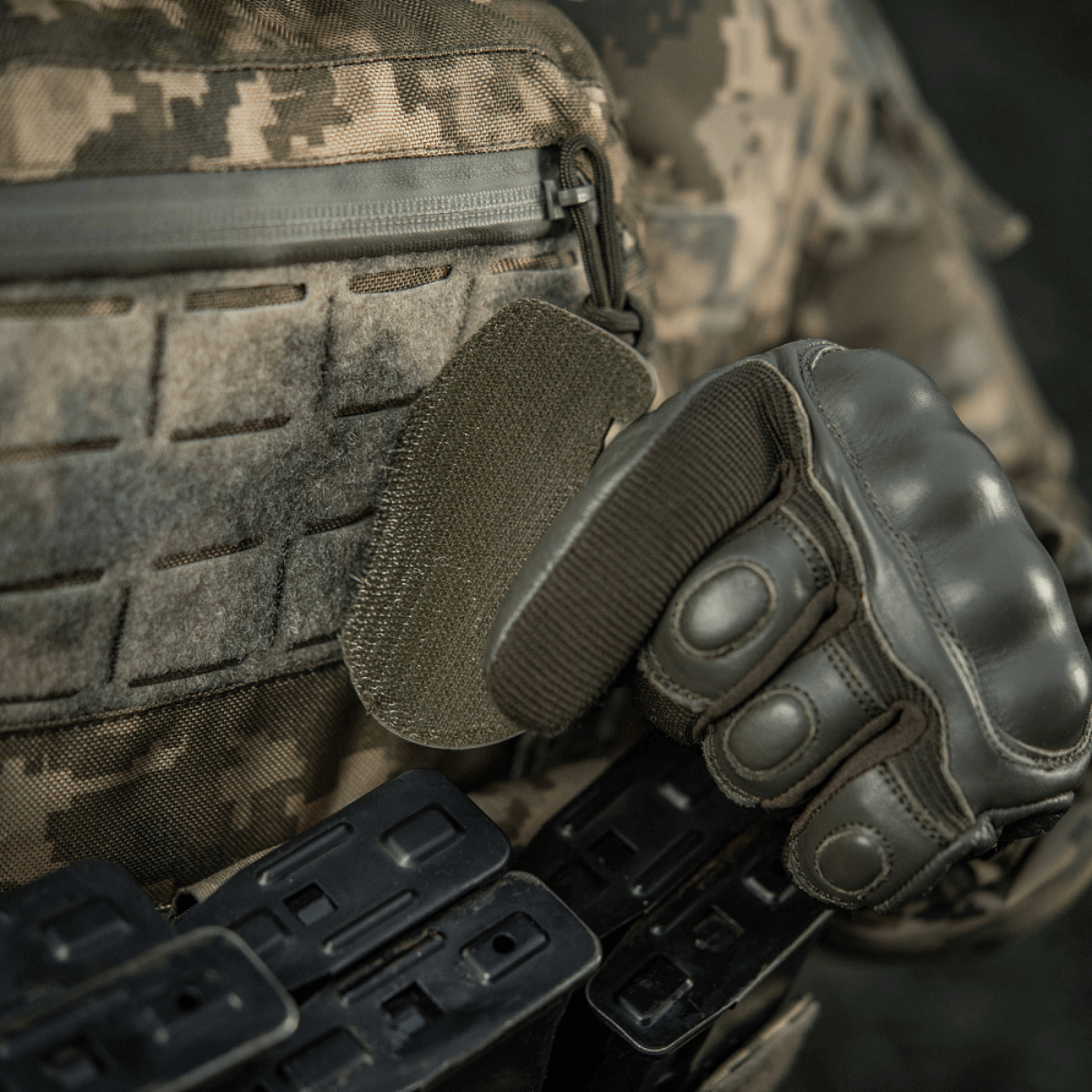 Sotilas taktisten varusteiden ja hansikkaiden kanssa kiinnittää M-Tac Patch M67 Kranaatti 3D PVC:n Velcro-takapuolella maastokuvioituun univormuun.