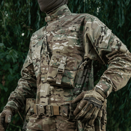 Henkilö maastopuvussa kantaa M-Tac PVS-14 -yönäkölaitteen laukkua olan yli ulkoilmataustalla.