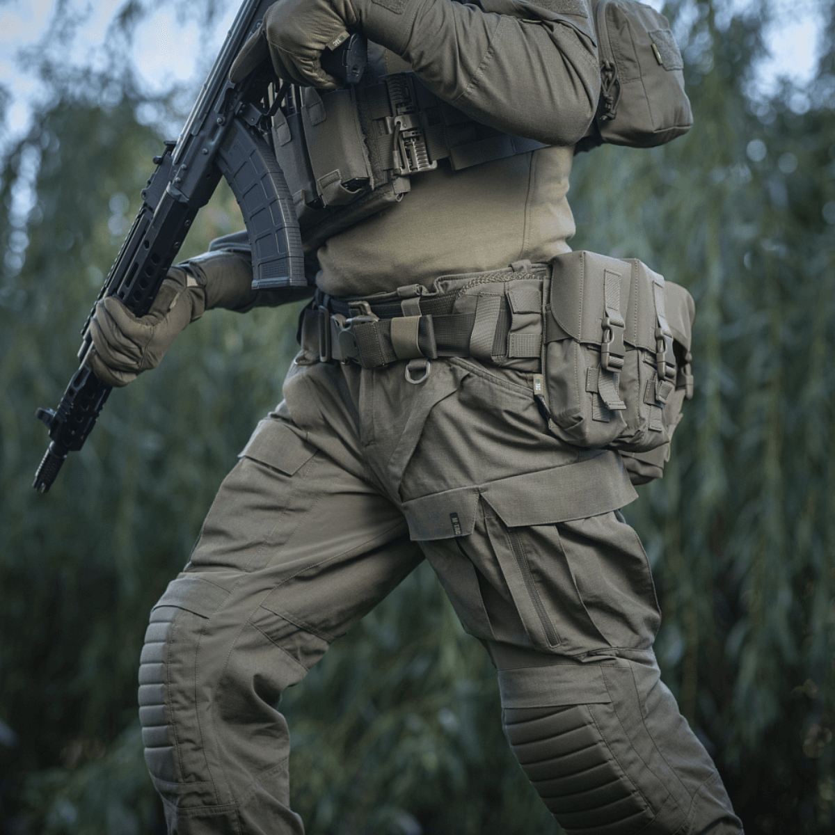 Henkilö pukeutuneena M-Tac War Belt ARMORiin ja taktiikka-varusteisiin luonnossa, valmis taistelutehtäviin.
