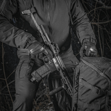 M-Tac Rygsæk Gen.II Elite Large, henkilö pitää asetta repun vieressä metsässä; repun kestävä muotoilu näkyy.