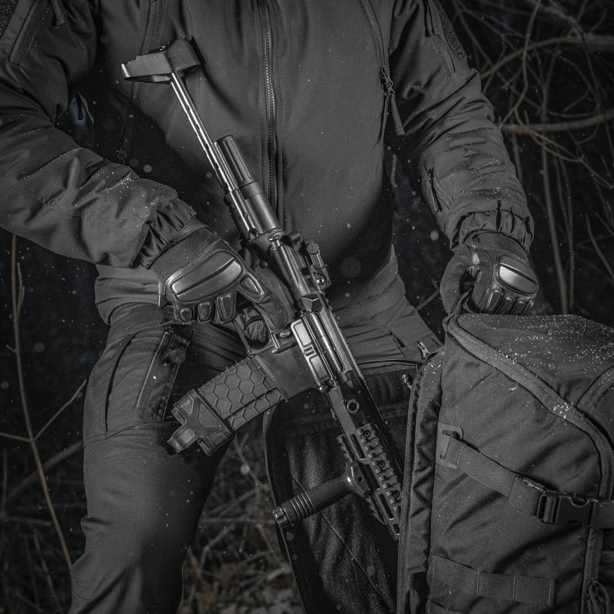 M-Tac Rygsæk Gen.II Elite Large, henkilö pitää asetta repun vieressä metsässä; repun kestävä muotoilu näkyy.