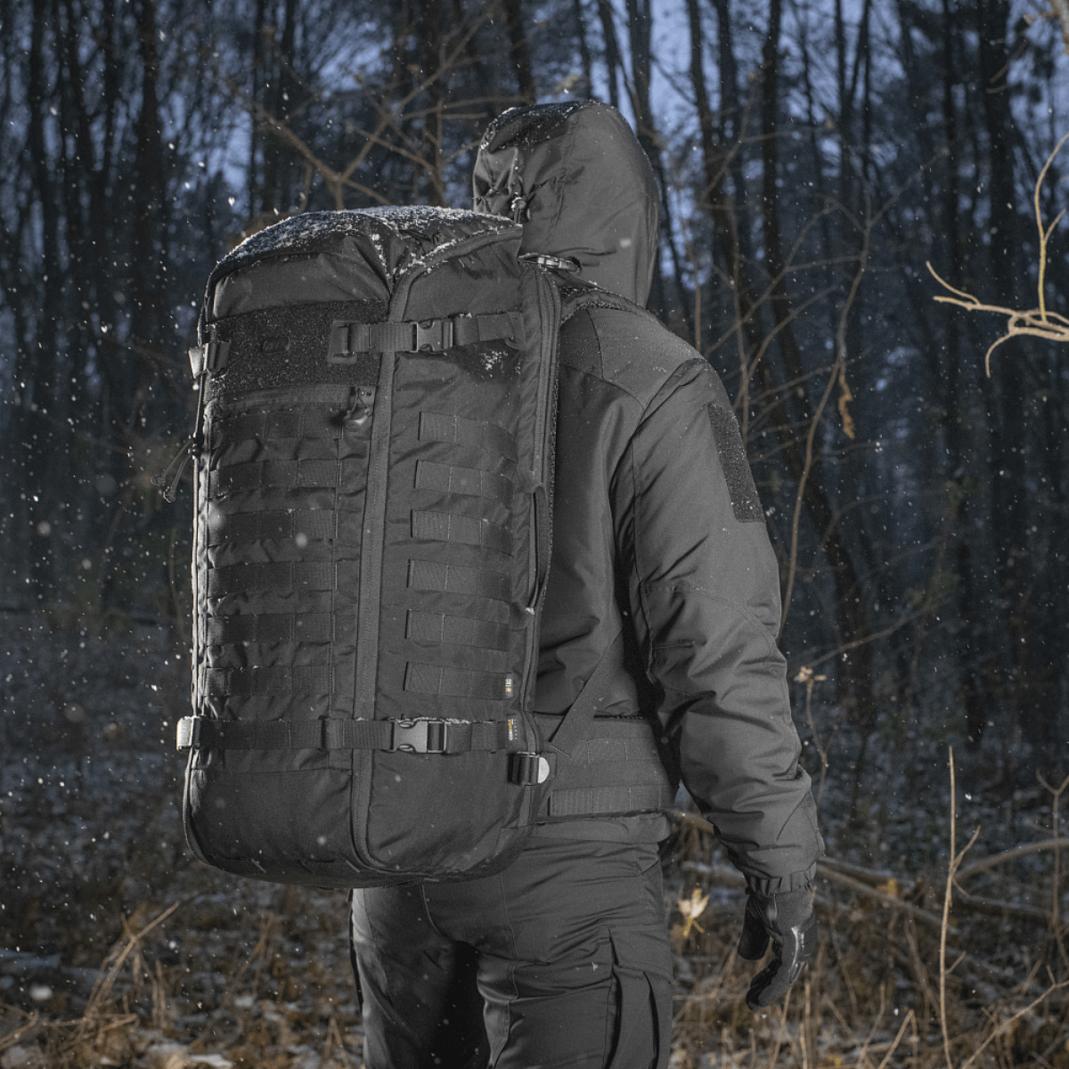 Henkilö kantaa M-Tac Rygsæk Gen.II Elite Largea talvisessa metsässä, lumisateen ympäröimänä.