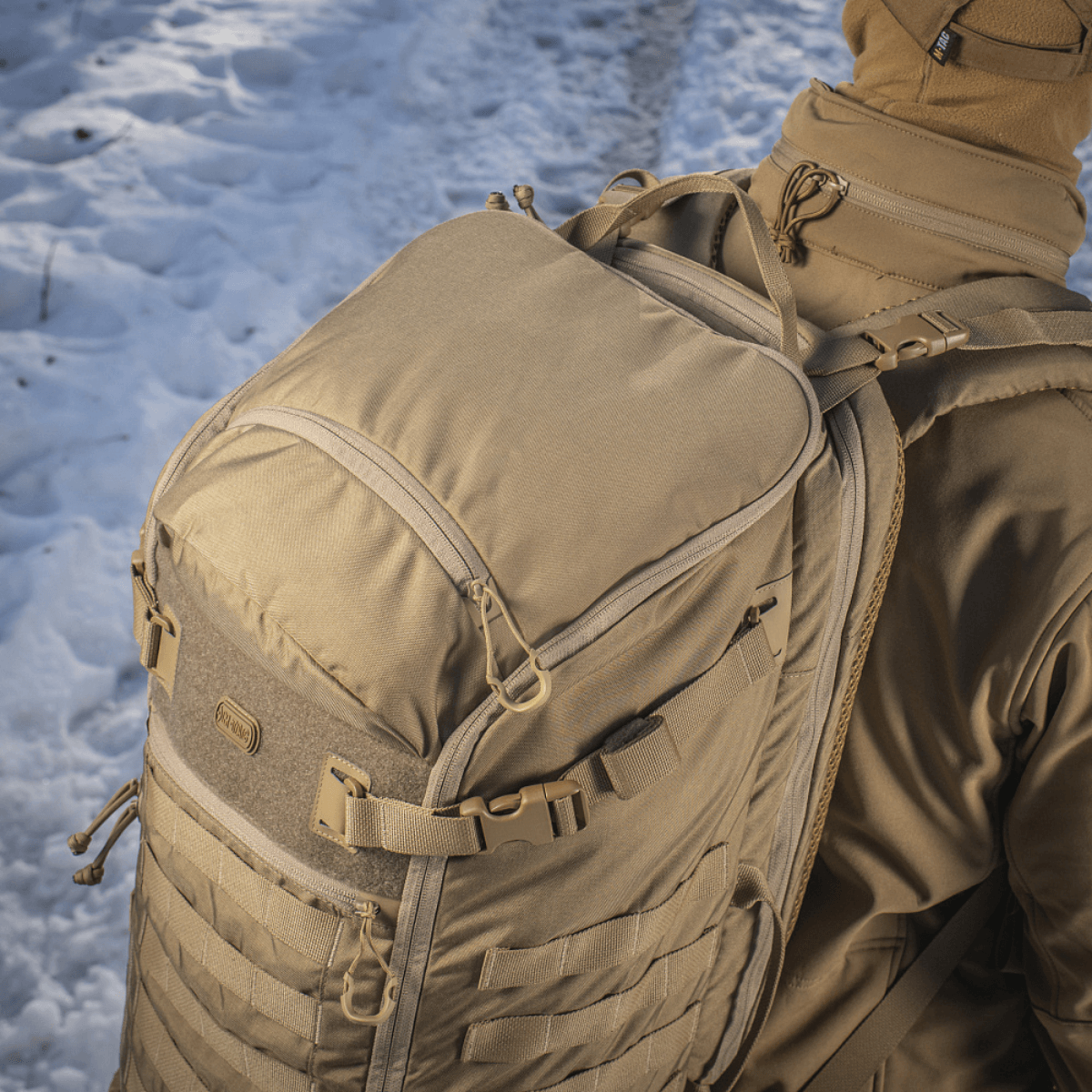 M-Tac Rygsæk Gen.II Elite Large, kannettu lumisessa maisemassa, esittelee Cordura 330D -materiaalin kestävyyttä, soveltuu aseiden kuljetukseen.