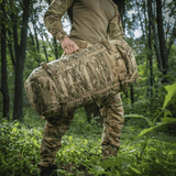 M-Tac Rygsæk Gen.II Elite Large, henkilö metsässä camokuvioisen repun kanssa, osoittaa kestävyyttä ja toiminnallisuutta ulkoilmassa.
