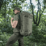 M-Tac Rygsæk Gen.II Elite Large, henkilö taktisissa vaatteissa metsäympäristössä, kuvaa repun monipuolisuutta ja kestävyyttä.