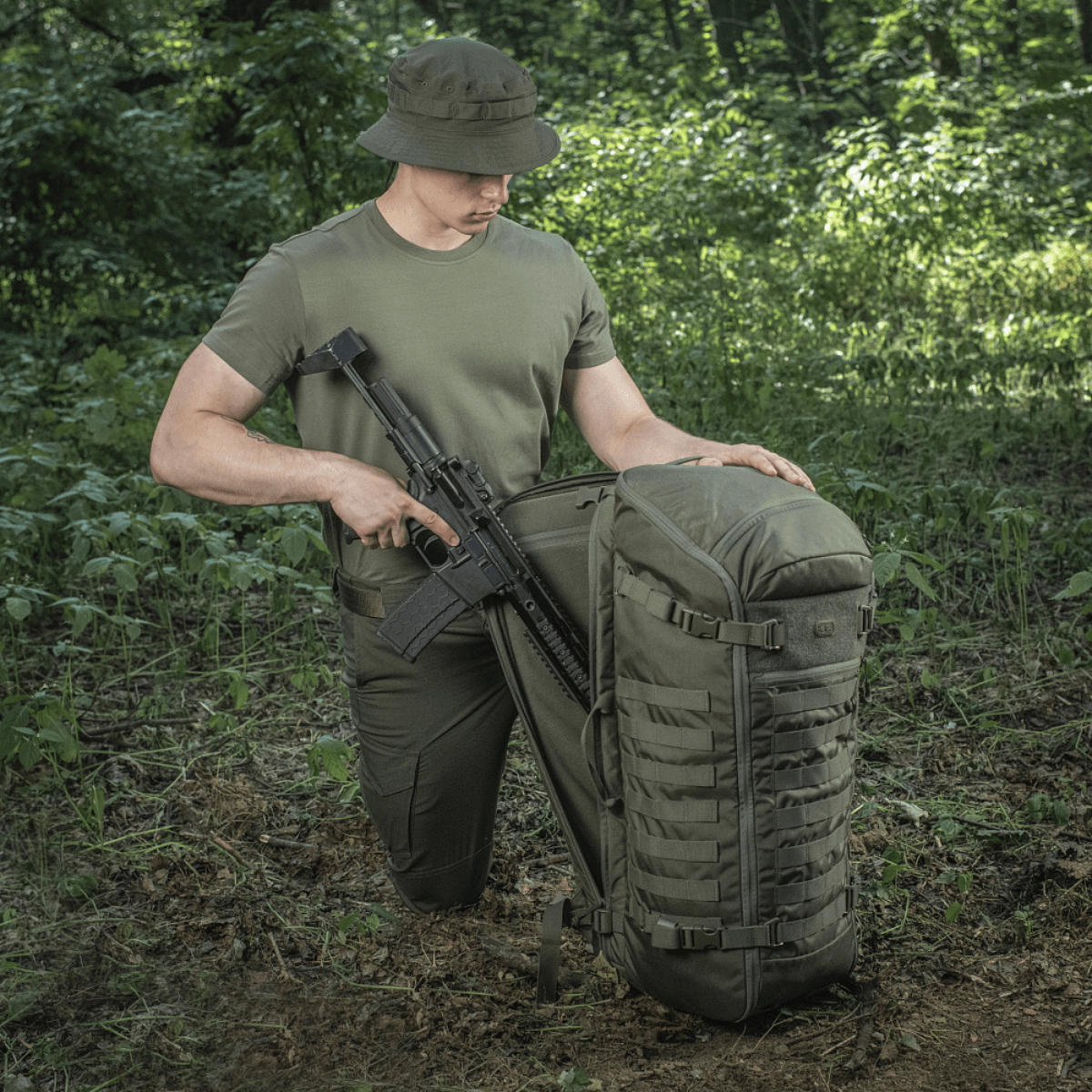 M-Tac Rygsæk Gen.II Elite Large, metsässä aseiden kanssa; näyttää kestävän rakenteen ja toiminnallisuuden ulkoiluseikkailuihin.