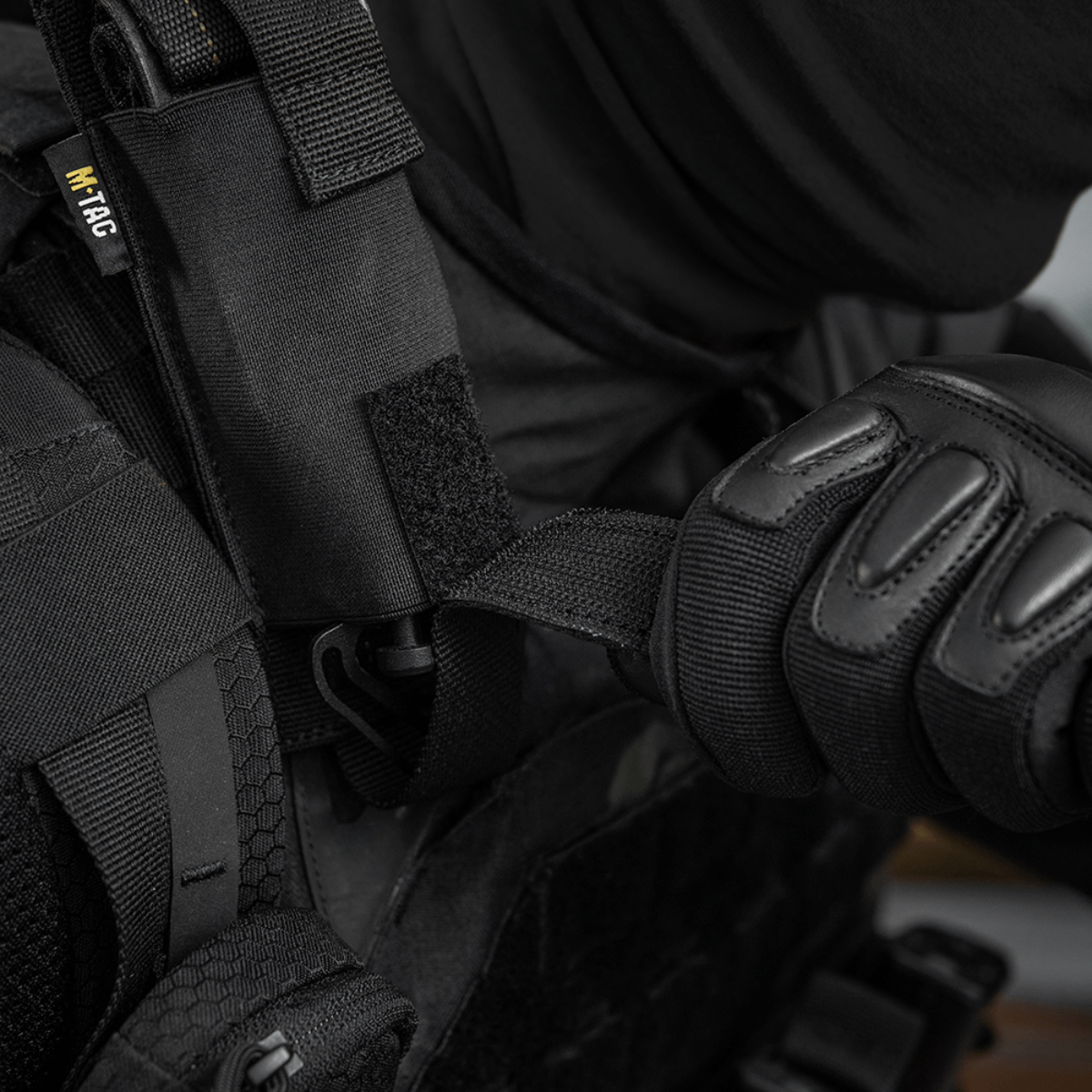Henkilö pukeutuneena mustaan suojavarustukseen käyttää M-Tac Tourniquet Pouch Elastisk med MOLLE Gen.II:tä kiinnitettynä liiviin.