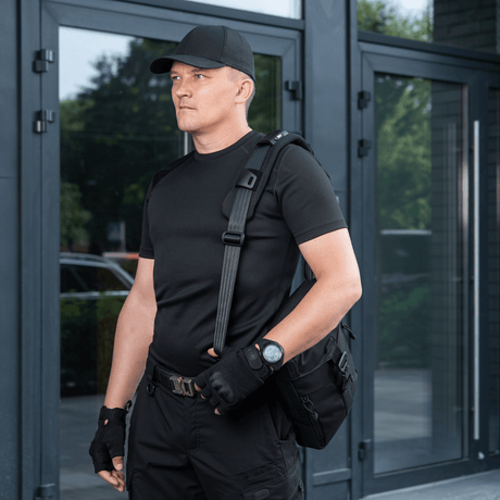 Mies kantaa laukkua M‑Tac olkahihnan pehmikkeellä 50 mm Elite lisämukavuutta ja tukea varten, pukeutuneena mustiin vaatteisiin modernin rakennuksen edessä.