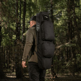 Mies kantaa M‑Tac Gun Backpack Case 105 cm Elite Hexiä metsässä, sopii AR-15- ja AK-kiväärien kuljetukseen. Cordura 500D -materiaali.