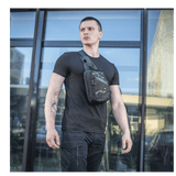 Mies M-Tac Sling Pistoltaske Elite Hex -laukulla lasirakennuksen edessä, näyttää monipuolisuutta ja tyyliä.