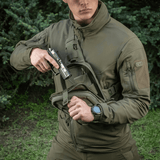 Henkilö, jolla on M-Tac Sling Pistoltaske Elite Hex, kantaa asetta metsätaustalla.