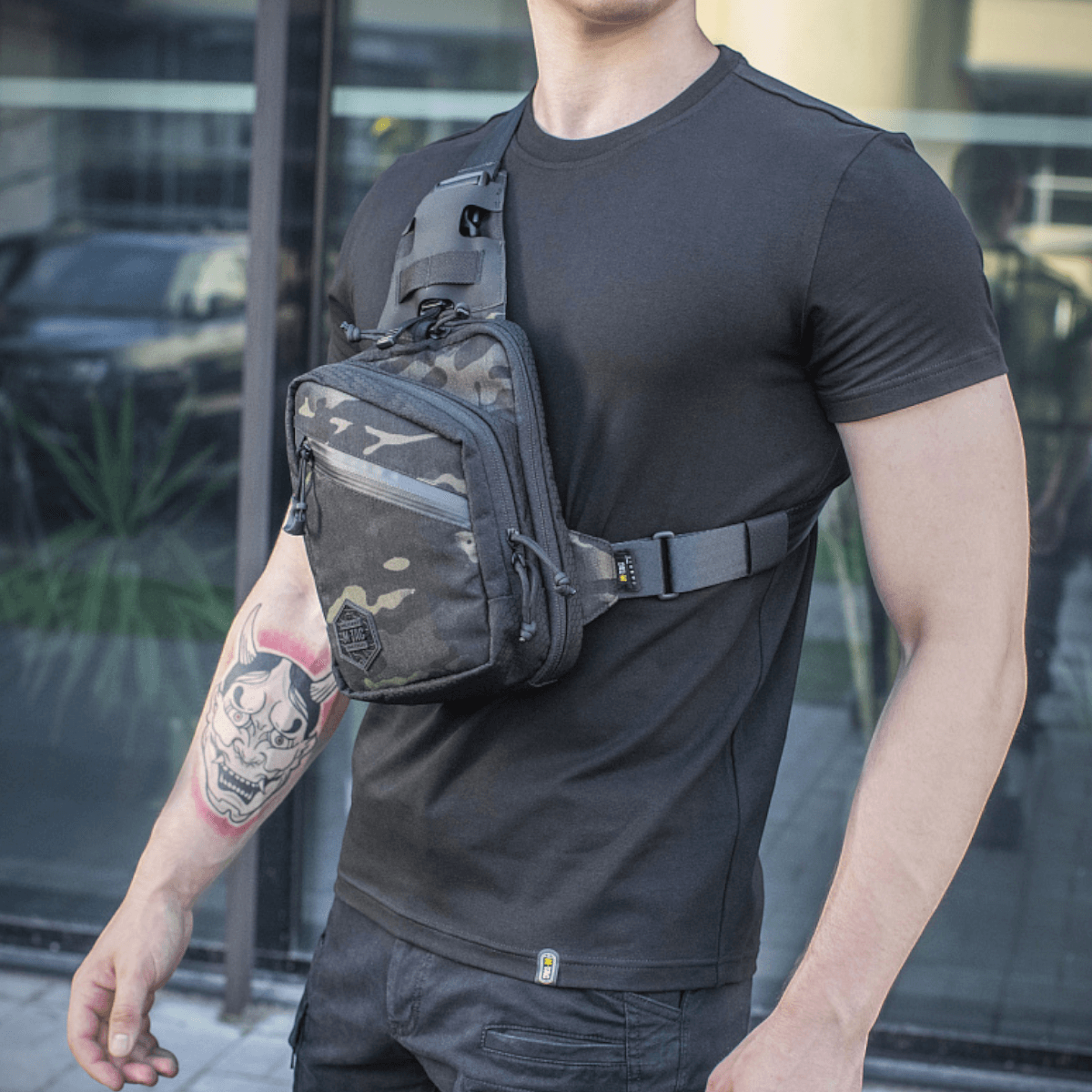 M-Tac Sling Pistoltaske Elite Hex miehen kantamana mustassa t-paidassa, näyttää laukun yksityiskohdat ja säädettävän olkahihnan.