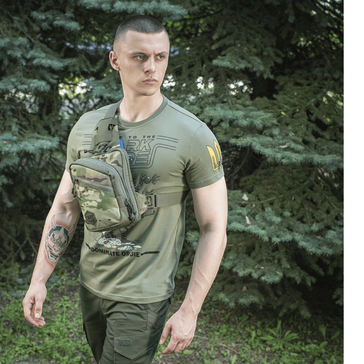 Mies vihreällä M-Tac Sling Pistoltaske Elite Hex -laukkulla, valmistettu vahvasta Cordurasta, säädettävä hihna metsäalueella.