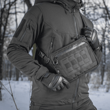Henkilö pukeutuneena mustaan takkiin M-Tac Admin-laukku Eliten kanssa, valmistettu kestävästä Cordura-materiaalista, lumisessa taustassa