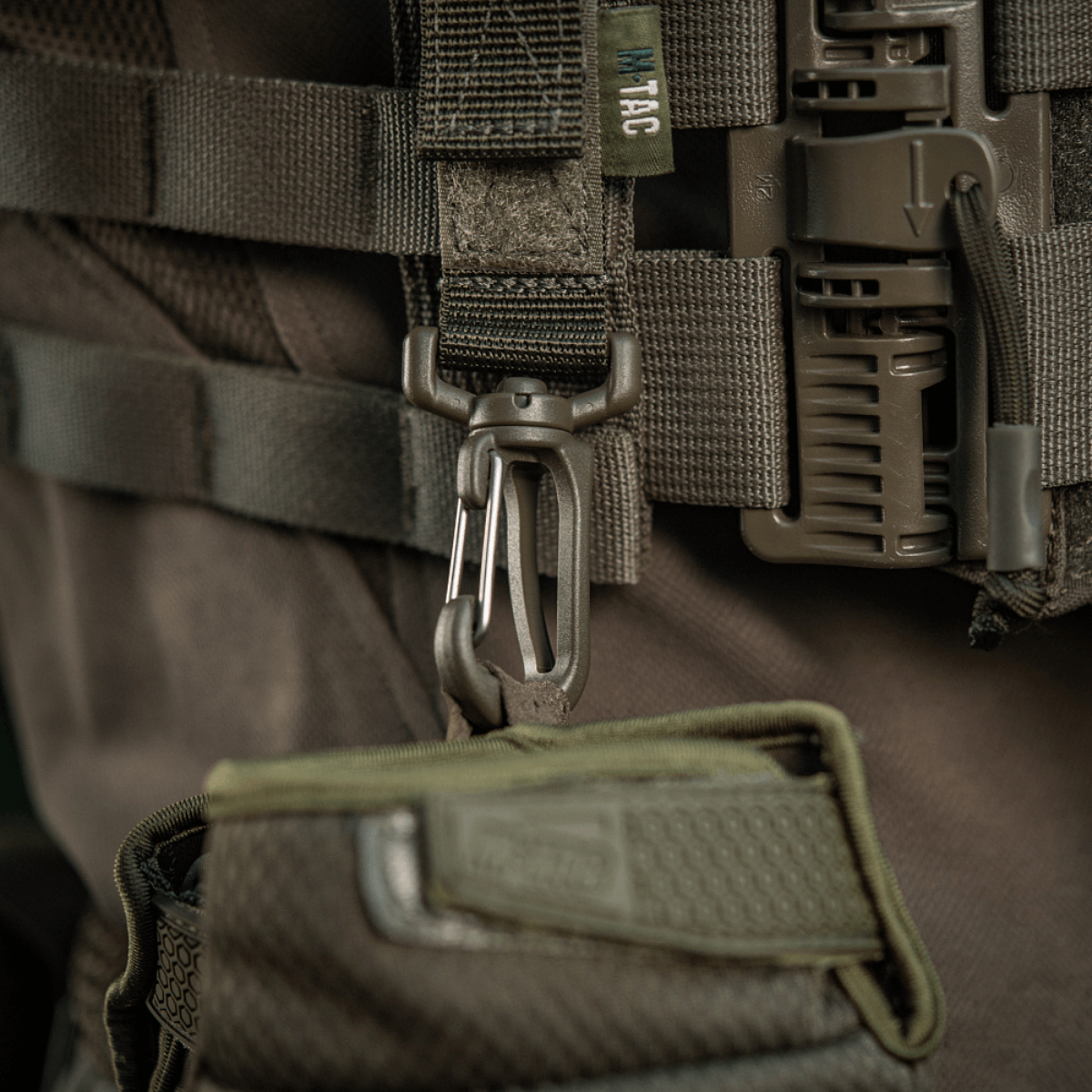 M-Tac karabiinihaka-lisävaruste kiinnitetty MOLLE-liiviin, säädettävä Velcro-remmi lisävarusteiden kiinnitykseen.