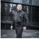 Mies M-Tac Cross Taske Slim Elite Hex -laukku olallaan, pukeutunut mustiin vaatteisiin modernin rakennuksen edessä.