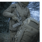 Henkilö kantaa asetta M-Tac repun vieressä luonnossa, esittäen repun kestävyyttä ja muotoilua.