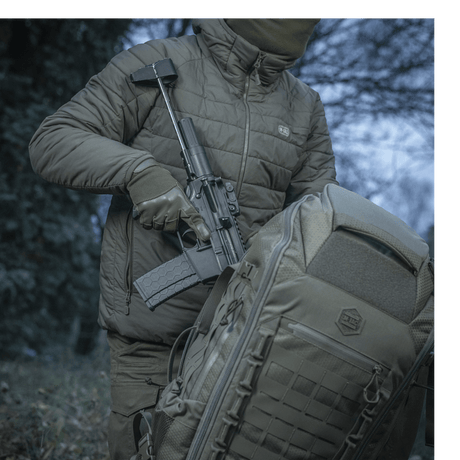 Henkilö kantaa asetta M-Tac repun vieressä luonnossa, esittäen repun kestävyyttä ja muotoilua.