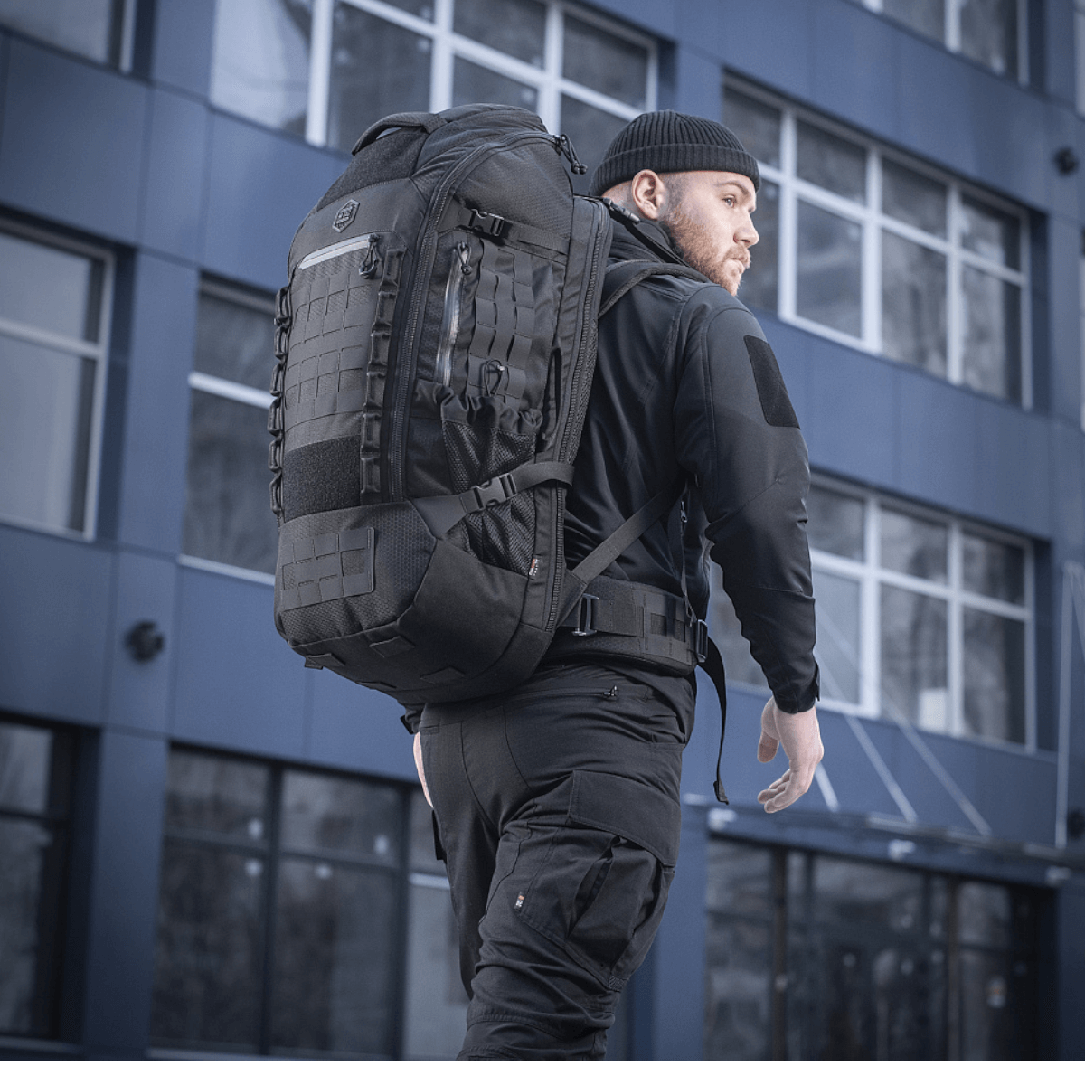 Mies kantaa M-Tac Rygsæk Large Elite Hex Gen. IV -reppua mustana, ergonomisesti suunniteltu toiminnallisuuteen ja kestävyyteen, nähty kaupunkiympäristössä.